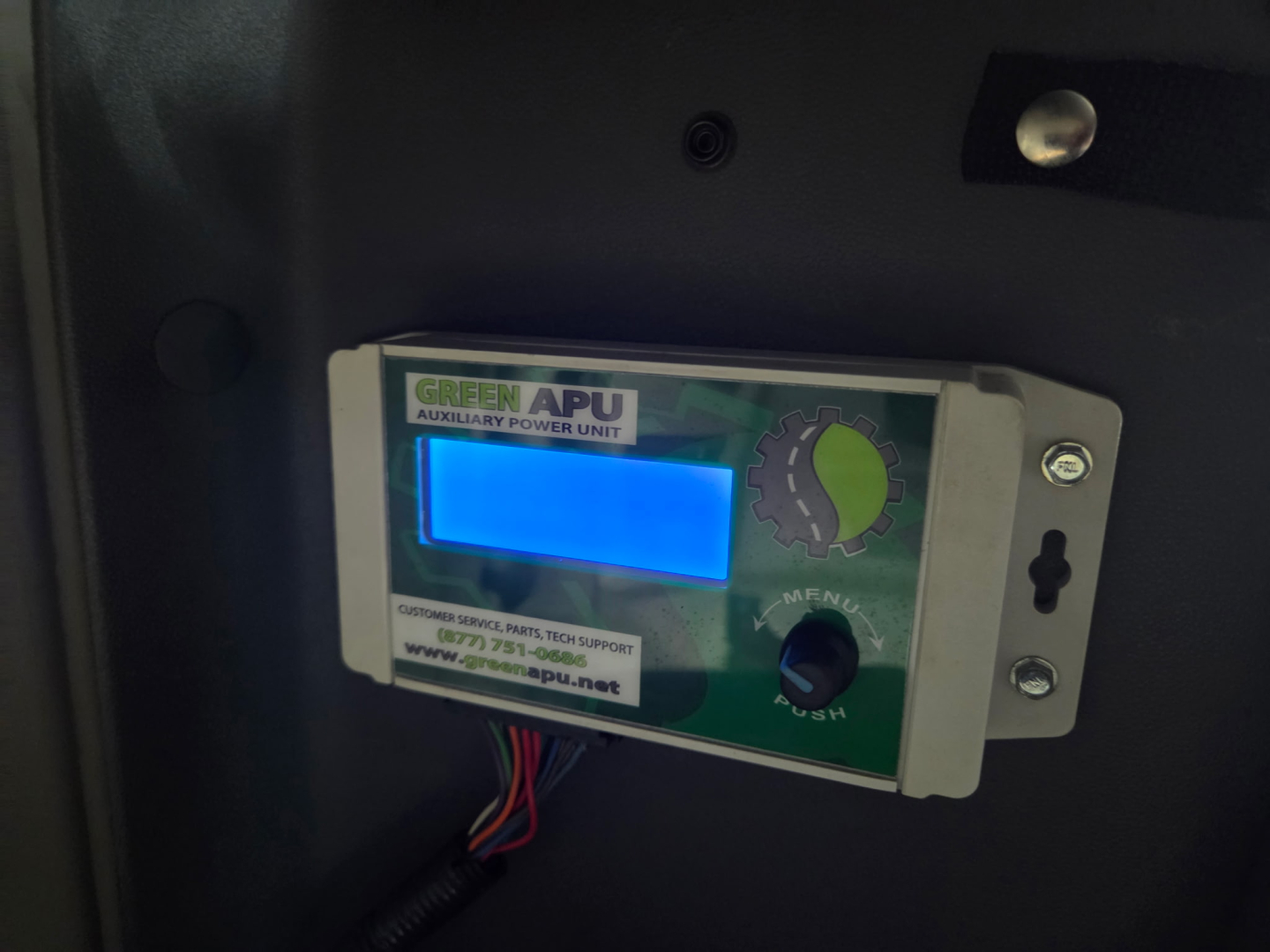 Truck cab interior showing Green APU auxiliary power unit controller panel with blue display and green logo, mounted inside dashboard.