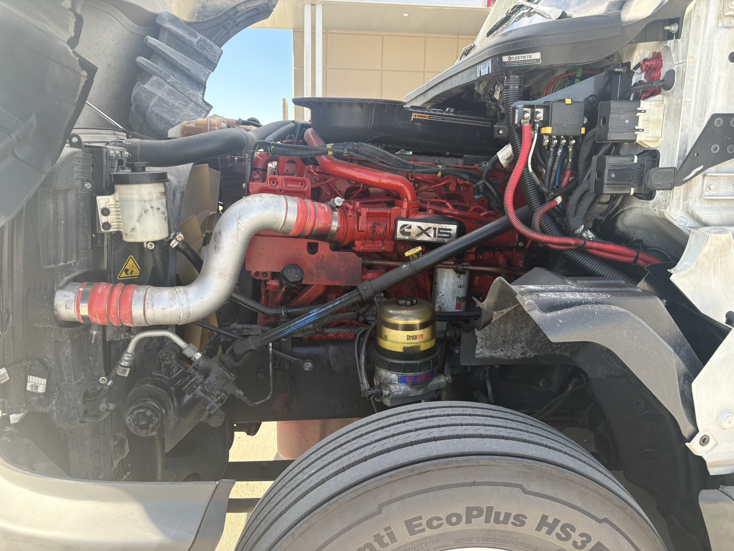 Truck engine bay showing Cummins-ISC inline engine with red components, intake manifold, hoses, and filter canister.