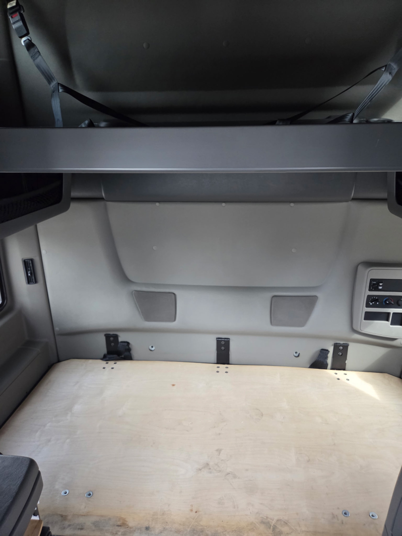 Truck sleeper cab cargo area showing flat wooden bed platform and rear wall with mounting brackets and side panels.