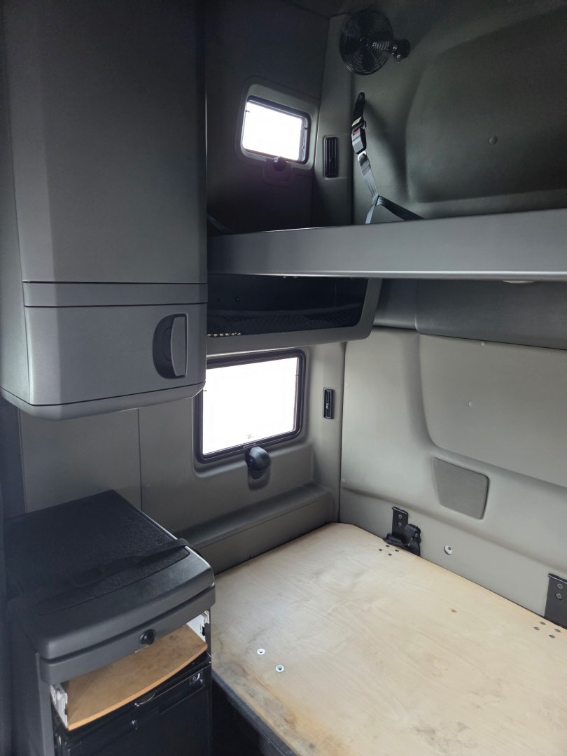 Truck cabin interior showing bunk bed area with window, overhead storage, and small desk area in a sleeper cab.