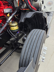 Truck engine bay with visible red engine components, black suspension, and a large front tire; yellow coolant/air filter reservoir and wiring harness visible.