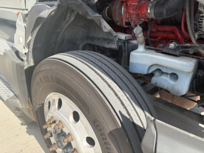 Truck front tire and wheel well with exhaust manifold and engine components visible; standard heavy-duty truck tire and wheel; part of an International LT625 cab-over? engine bay area visible.