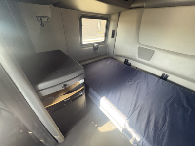 Interior sleeper cab of a 2023 International LT625 showing a single sleeper berth with blue mattress, window, storage cabinet, and compact fridge.