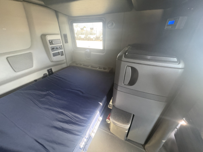 Interior of a sleeper truck cab with a bed on the left, a small cabinet and refrigerator on the right, window in the middle, and control panels along the walls.