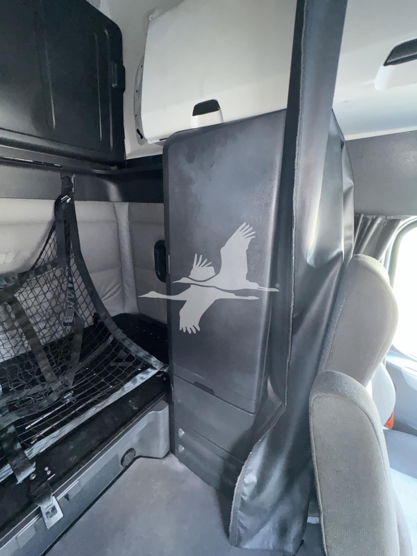 Truck interior showing sleeper cab partition with Freightliner logo on black panel, storage shelf and netting visible in rear area.