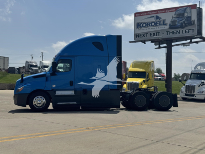 Freightliner Cascadia 126 sleeper tractor in blue with white bird graphic on door, parked in lot beside billboard; truck trailer connection area visible