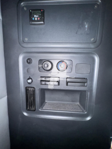 Dashboard panel with switches and circular dials and a small storage pocket in a Freightliner Cascadia cab interior. Visible are two round knobs, a red/blue climate control ring, a few toggle switches, and a small empty tray.