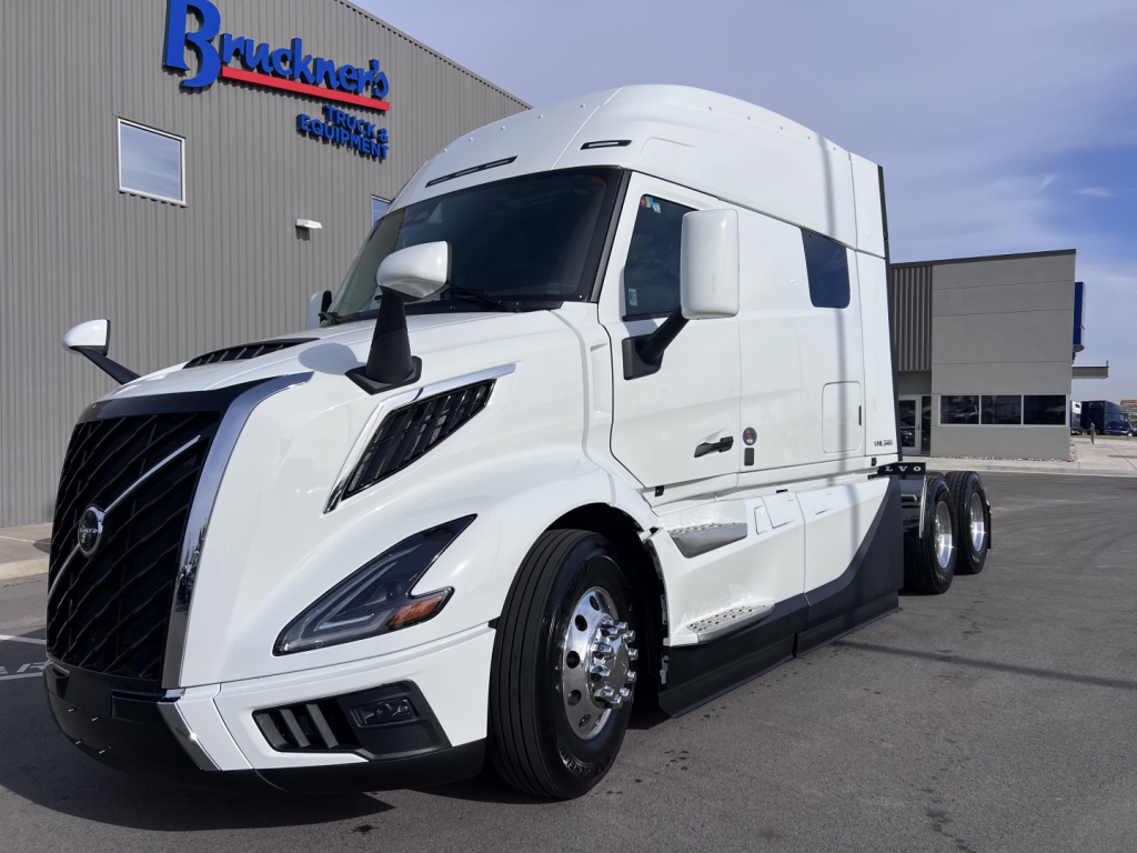 Truck engine bay not visible; a white Volvo VNL64T840 sleeper cab tractor parked outside dealership; side view shows triple rear axles and aerodynamic front with large grille.