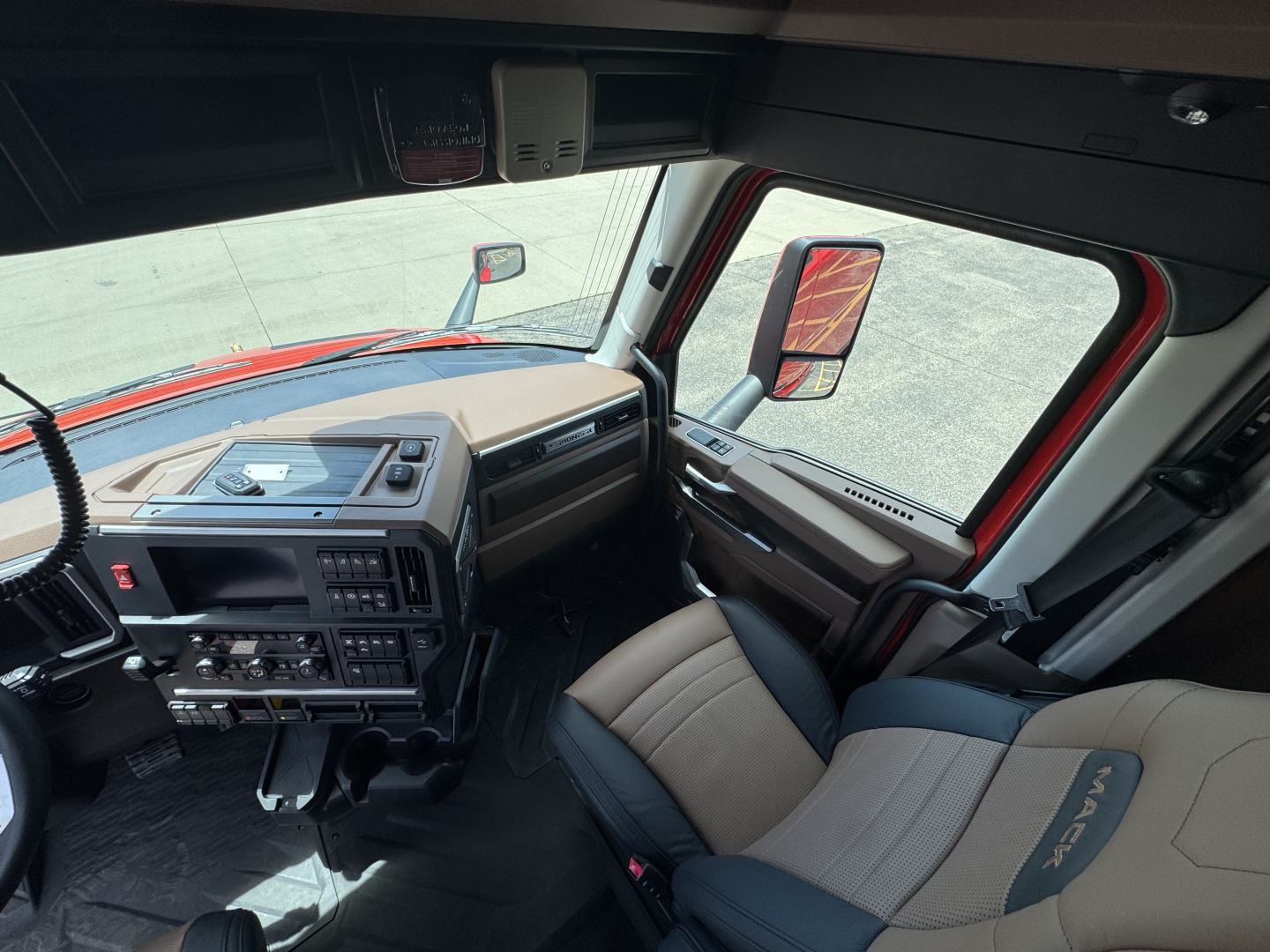 Cab interior of a new 2027 Mack PR64T76H truck showing dashboard and driver seat area with steering wheel, controls, and red exterior mirror.