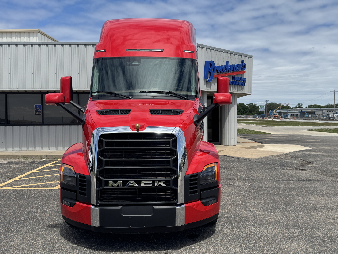 Front view of a red Mack semi-truck with large black grille, chrome accents, and 'MACK' prominently on the grille. Side mirrors visible, truck is in a parking lot.