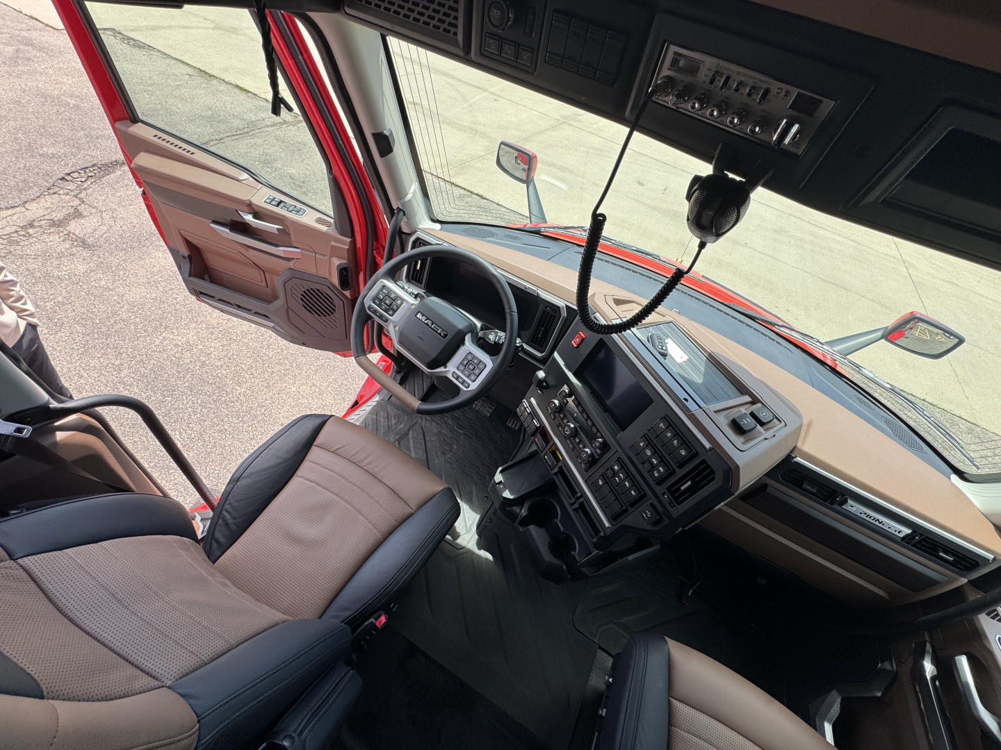 Mack PR64T76H truck cab interior showing steering wheel, dashboard, and control panel from driver’s seat; tan and black seats; windshield and side mirrors visible.