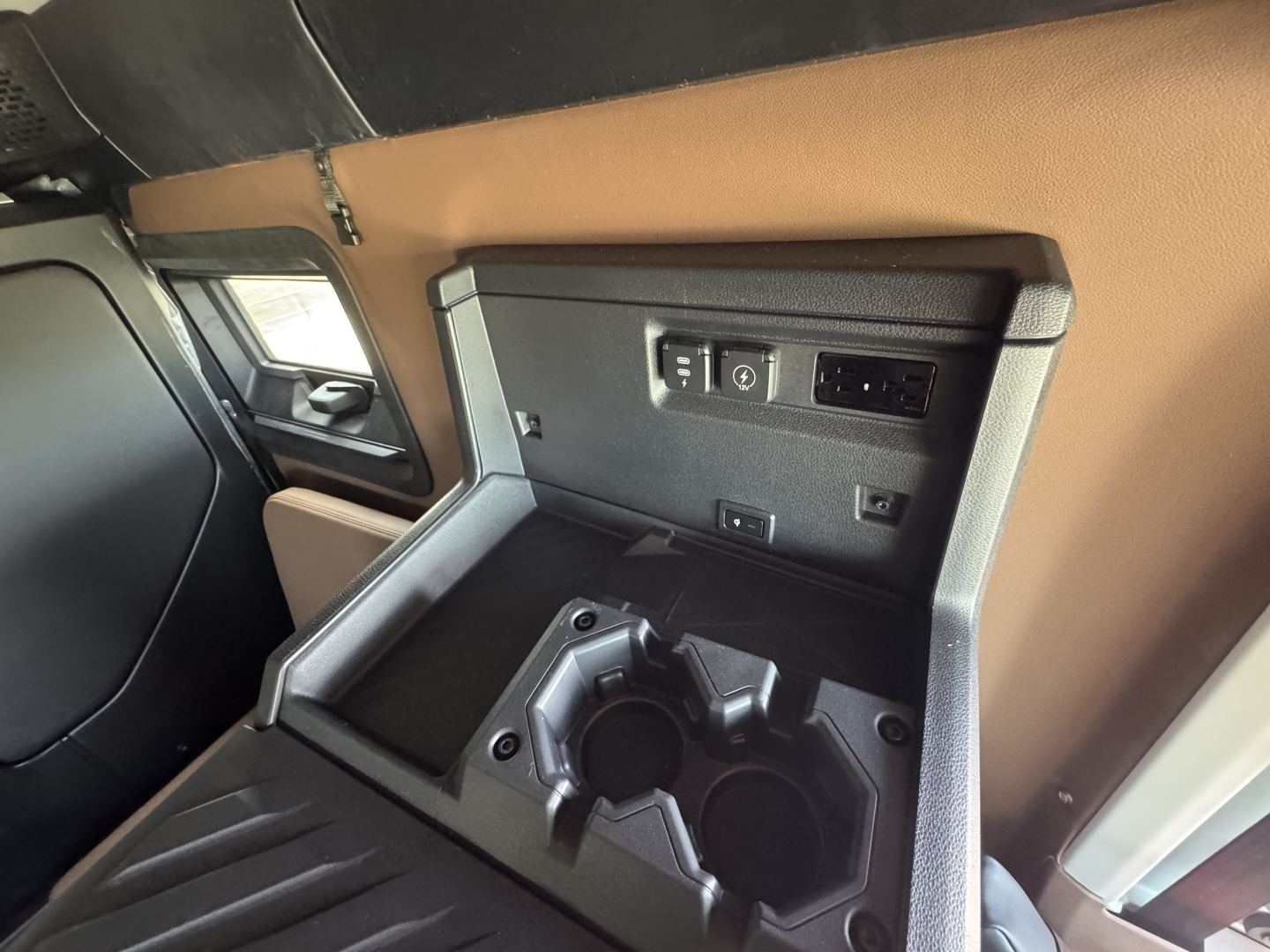 Truck interior: 12V power outlet panel with two USB ports, a standard AC outlet, and cup holders in the center console area; brown/black dashboard and side panels.