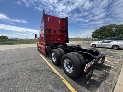 Truck chassis with red sleeper cab, rear view shows dual rear axles, fuel tanks, and wheel rims. Clear sky, parking lot. Visible on bumper: Mack PR64T76H model details not legible.