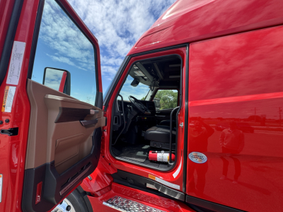 Truck cab interior visible through open door of red Mack PR64T76H. Dashboard and steering wheel seen; exterior door area with side mirror and badge.