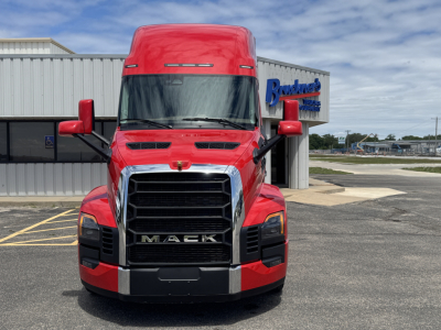 Front view of a red Mack semi-truck with large black grille, chrome accents, and 'MACK' prominently on the grille. Side mirrors visible, truck is in a parking lot.