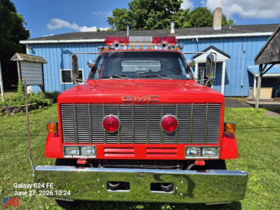 GMC Topkick 7000 pumper fire truck front view with chrome grille, red body, and dual red headlights; blue building in background.