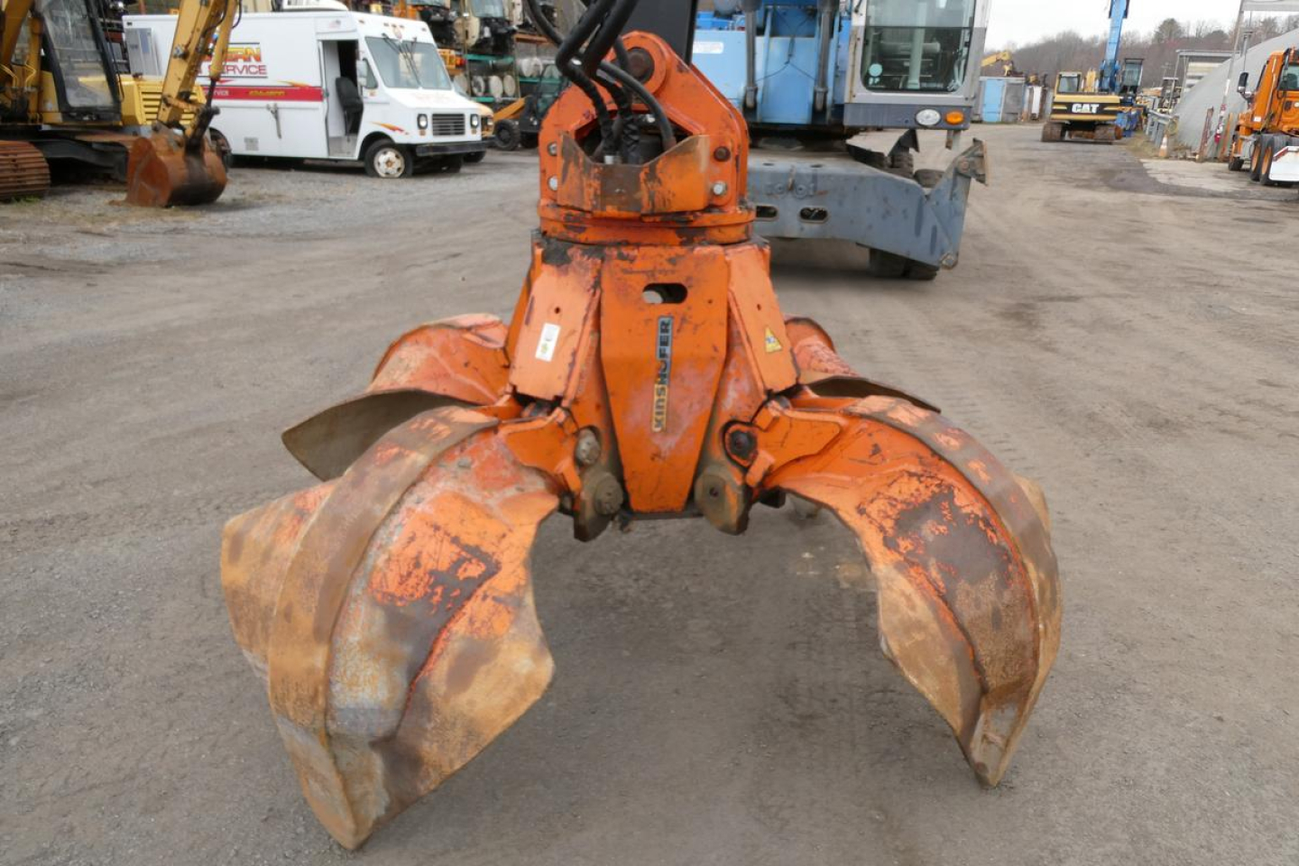 Orange Terex MHL340D grapple attachment seen from front, two large curved claw buckets and hydraulic links; industrial yard in background.