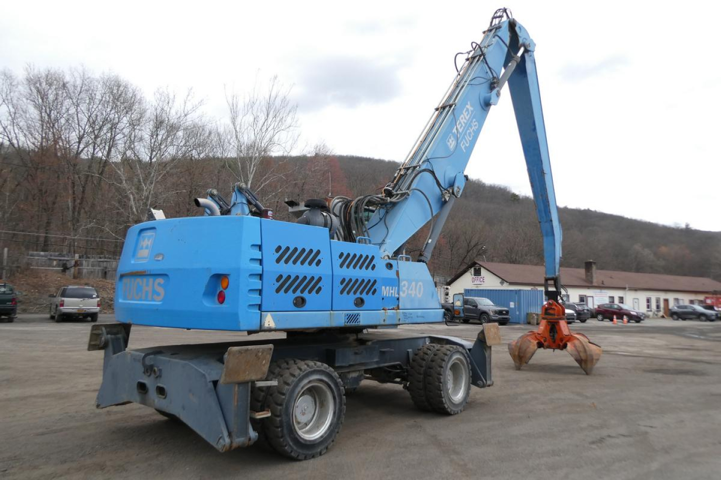 Terex MHL340D material handler crane in blue showing hydraulic arm and grapple, rear view with tires and operator cab, recycling equipment on site
