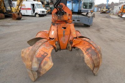 Orange Terex MHL340D grapple attachment seen from front, two large curved claw buckets and hydraulic links; industrial yard in background.