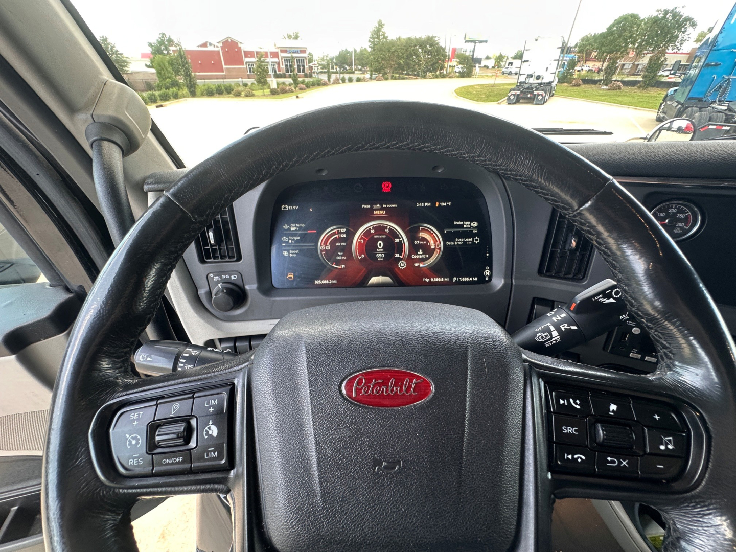 Peterbilt 579 truck dashboard and steering wheel with digital gauge cluster visible, center emblem shows Peterbilt logo, steering wheel controls present, lightly tinted windshield view outside.