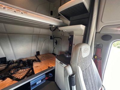 Peterbilt 579 truck cab interior showing sleeper area with storage shelf, overhead compartments, and passenger seat; cargo area with wooden platform and tie-downs.