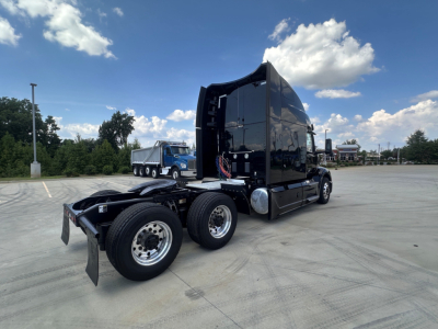 Peterbilt 579 tractor rear view showing sleeper cab and dual rear axles, black exterior, chrome fuel tanks, with clear sky and parking lot in background.