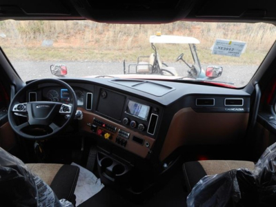Cab interior of a new Freightliner Cascadia 126, viewing the dashboard, steering wheel, and center console with brown trim.