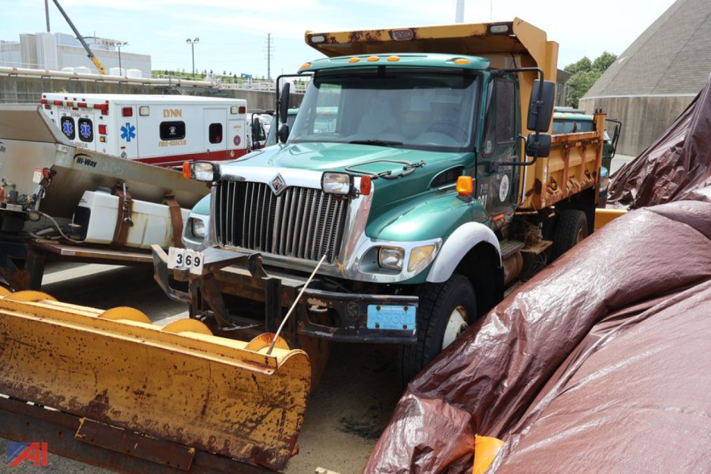 Dump truck front view showing 2007 International WorkStar 7400 with green cab and yellow bed, snow plow attachment, and surrounding equipment.