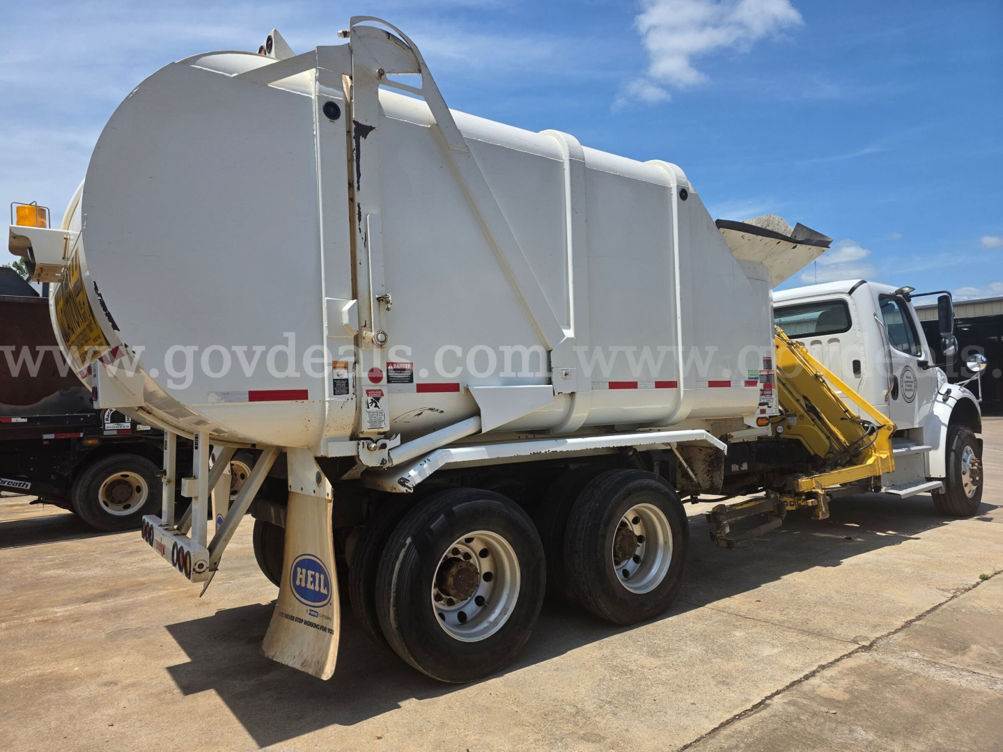 Waste collection truck with a large white tank mounted on a chassis, rear view shows dual rear axles and a hydraulic mechanism; visible labels and stickers on tank and mudflaps.