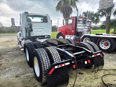 Truck chassis and cab viewed from rear with dual rear axles, International LT625 daycab, Cummins engine, heavy-duty tires, red and gray trucks in background.
