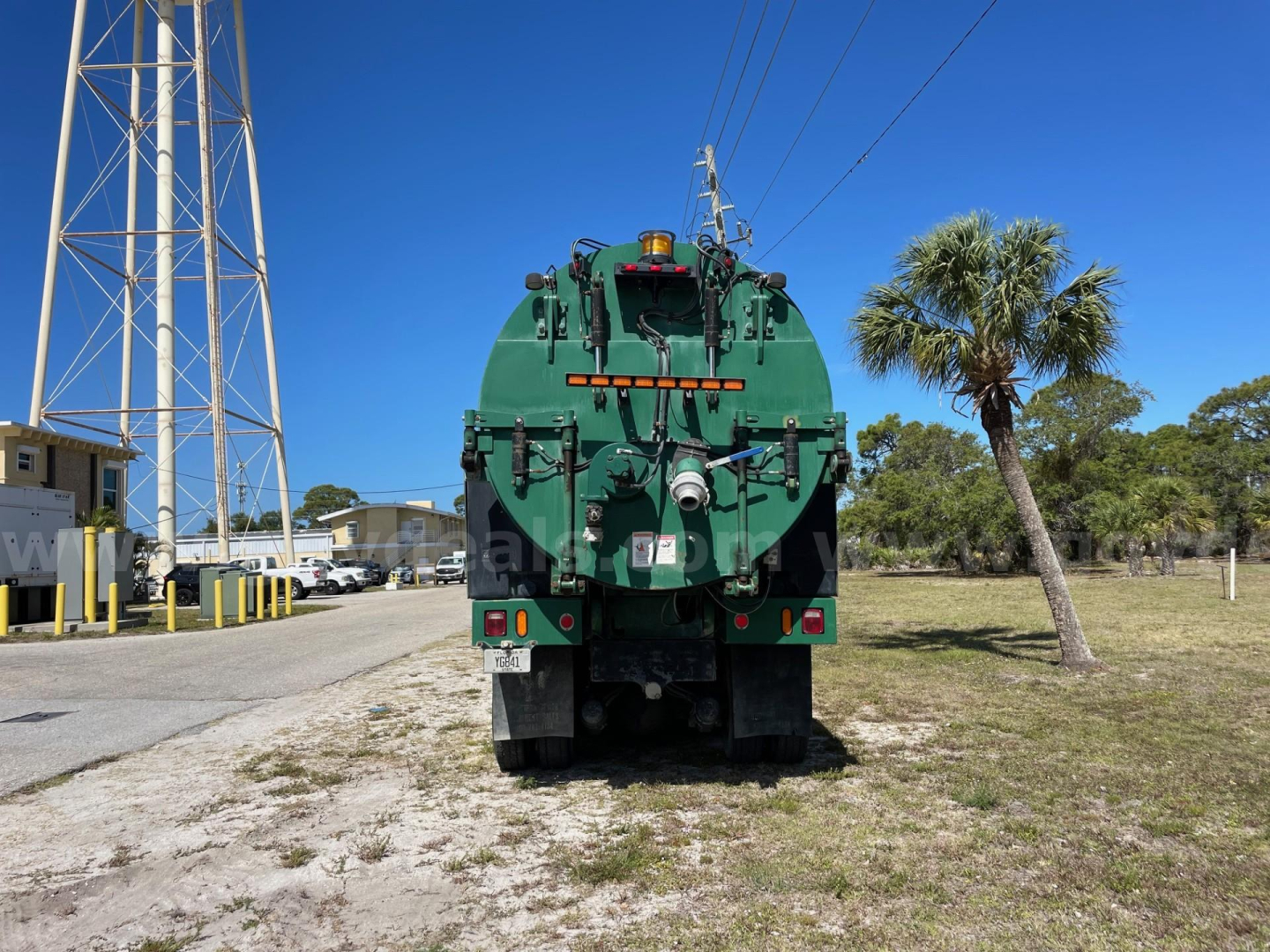 Rear view of a green Freightliner vacuum tanker truck (2013 Freightliner 114SD) with rear hoses and equipment, palm tree, and clear blue sky.