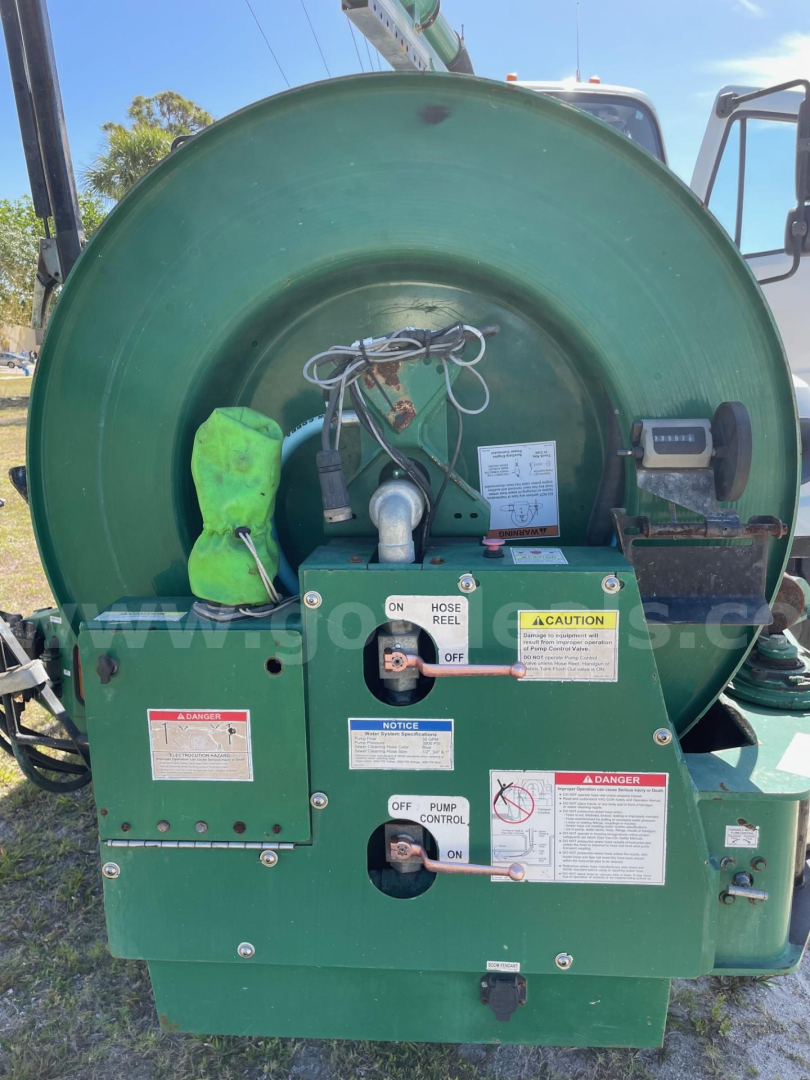 Green Freightliner 114SD waste vacuum truck rear machinery with hoses, valves, warning labels, and control switches.
