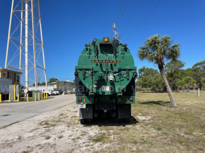 Rear view of a green Freightliner vacuum tanker truck (2013 Freightliner 114SD) with rear hoses and equipment, palm tree, and clear blue sky.
