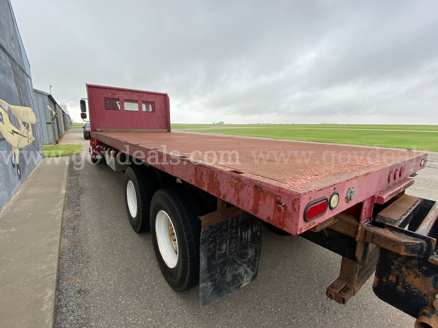 Flatbed trailer on a Freightliner M2 106 chassis showing dual rear wheels and rusty flatbed with black mud flaps.