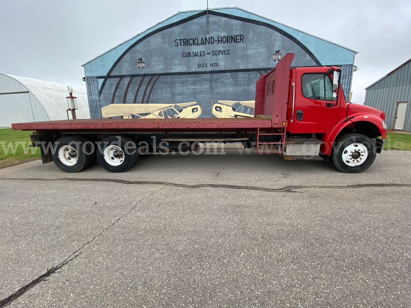 Flatbed truck with red cab and long flatbed, Freightliner M2 106, side view showing triple rear axles.