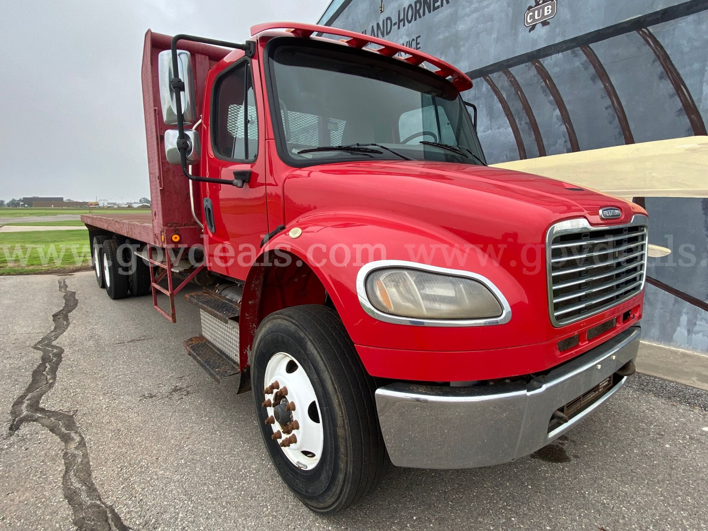 Freightliner M2 106 flatbed truck in red, front-left view showing cab, grille, headlight, and flatbed cargo area.