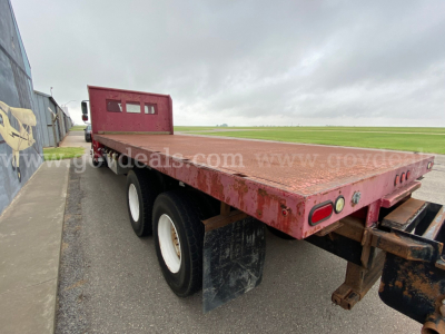Flatbed trailer on a Freightliner M2 106 chassis showing dual rear wheels and rusty flatbed with black mud flaps.