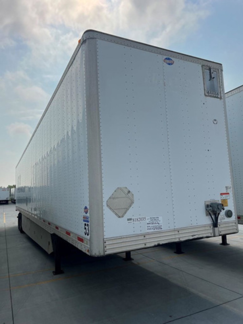 Trailer van rear view showing aluminum riveted box trailer with rear doors and bumper, storage decals near bottom, wheel chocks, and support legs.