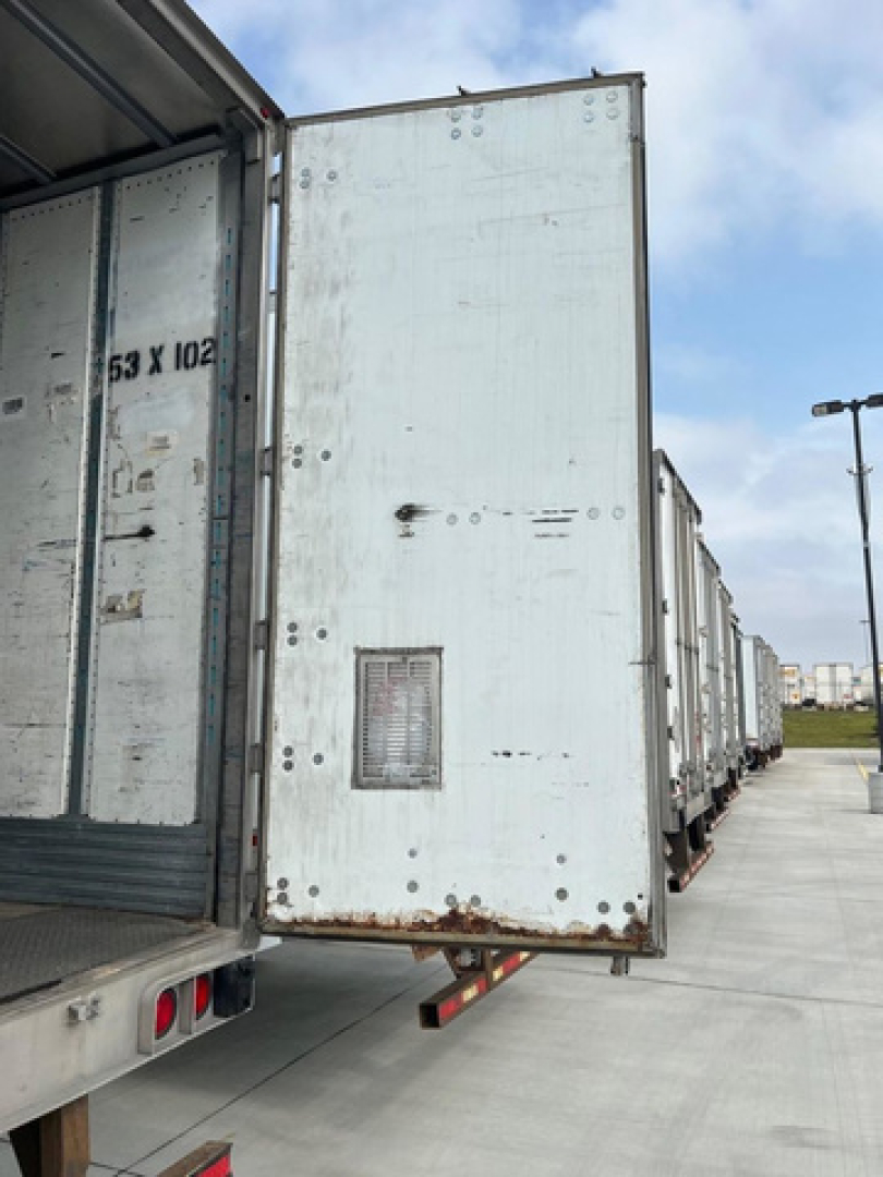Utility van trailer rear door open showing worn white exterior, hinge area, and rock-steady support legs; side view of row of trailers in lot.