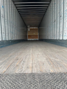 Trailer interior showing empty cargo space with wooden floor, ribbed metal walls, and open rear; no visible text or labels.