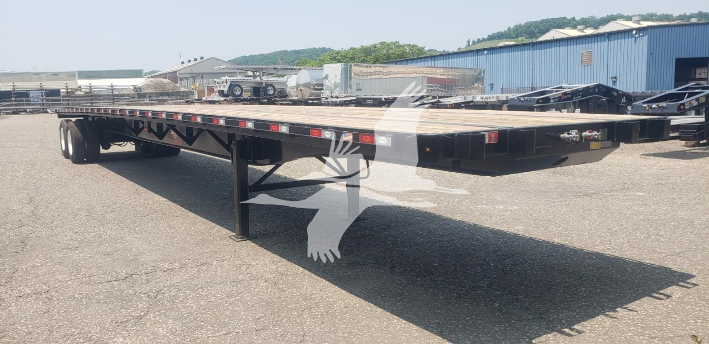 Flatbed trailer with black frame and tandem axles showcased in yard; long low-profile deck, side rails, and support legs visible.