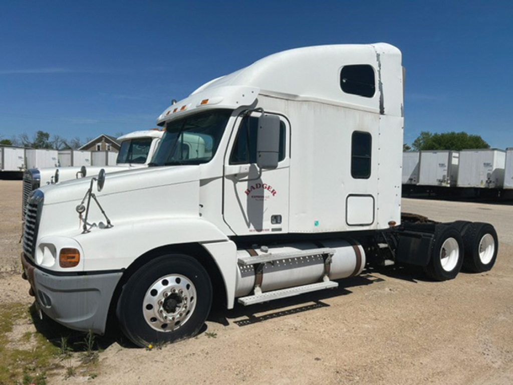 White Freightliner Century Class C120 tractor in a yard, with sleeper cab and dual rear axles; visible text includes 'FREIGHTLINER' emblem on door, and side mirrors and steps; no other legible labels.