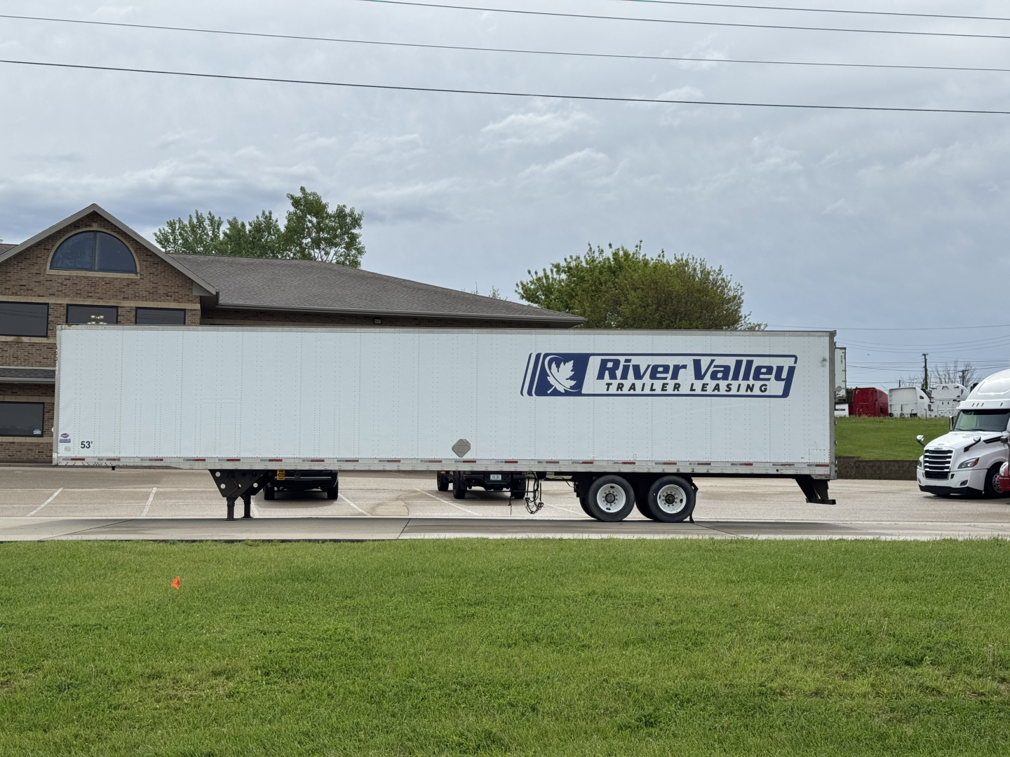 Trailer side view showing River Valley Trailer Leasing logo on a long white utility trailer attached to a truck, with a dealership setting and a building in the background.