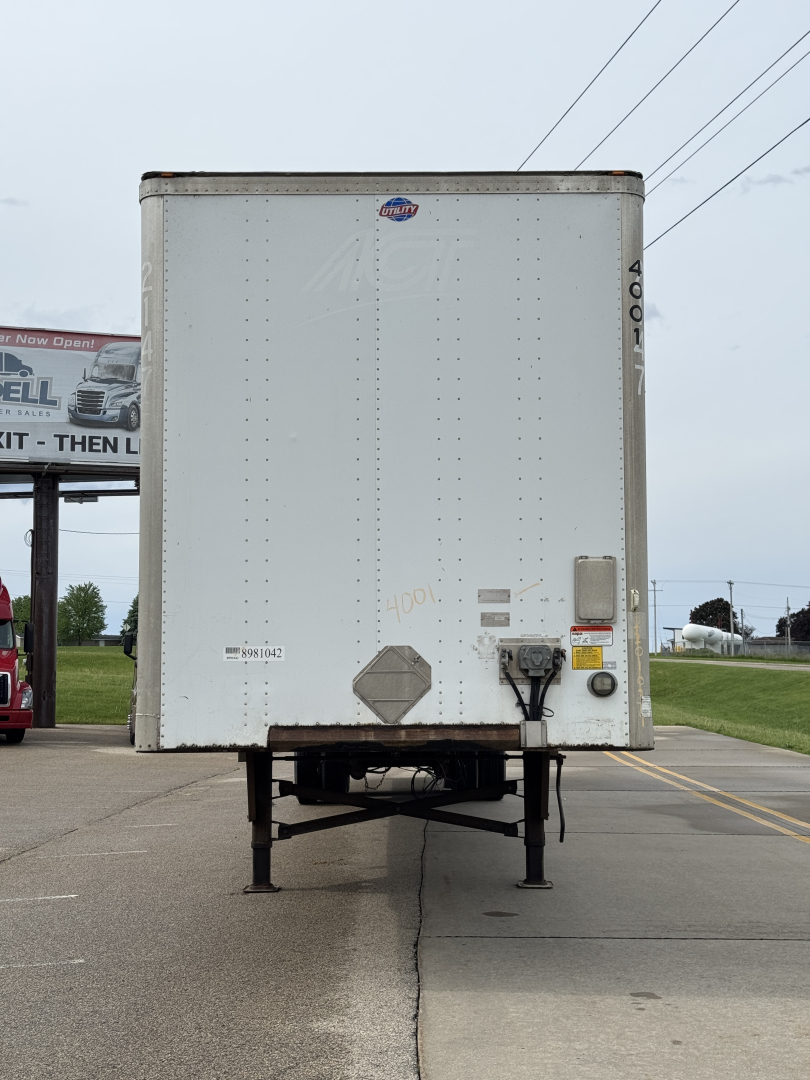 Trailer back view showing plain white cargo box with support legs, small diamond placard, and rear lights; looks like a utility trailer attached to a truck lot.