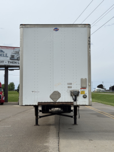 Trailer back view showing plain white cargo box with support legs, small diamond placard, and rear lights; looks like a utility trailer attached to a truck lot.