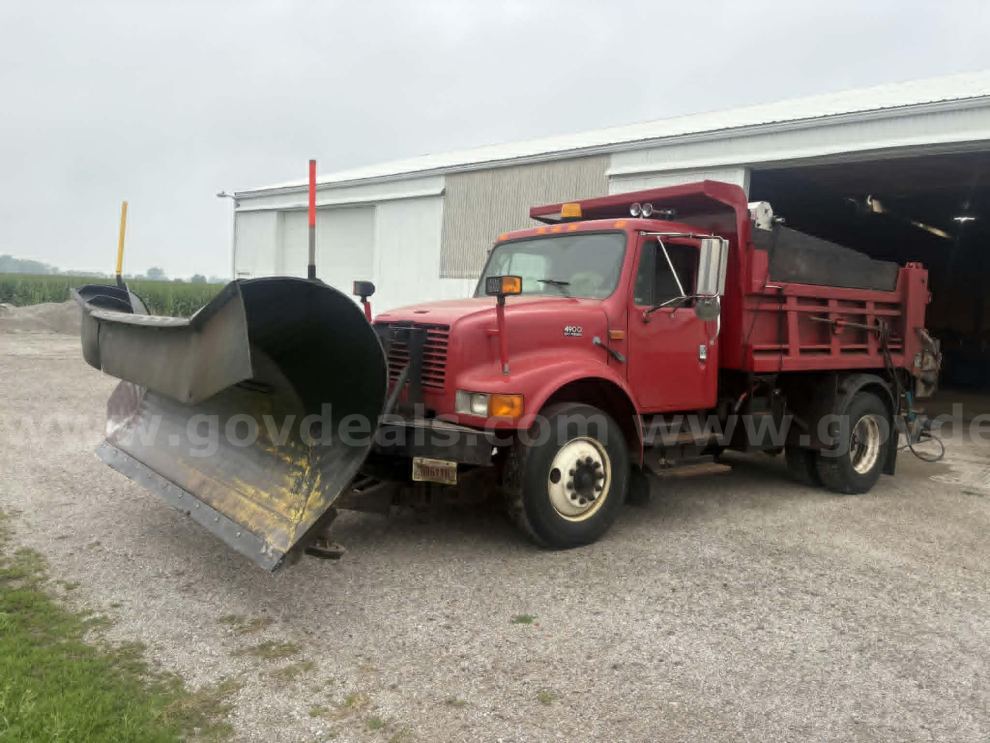 2000 International 4900 dump truck with snowplow or front plow attachment, red cab and dump bed, parked outside a large shed.