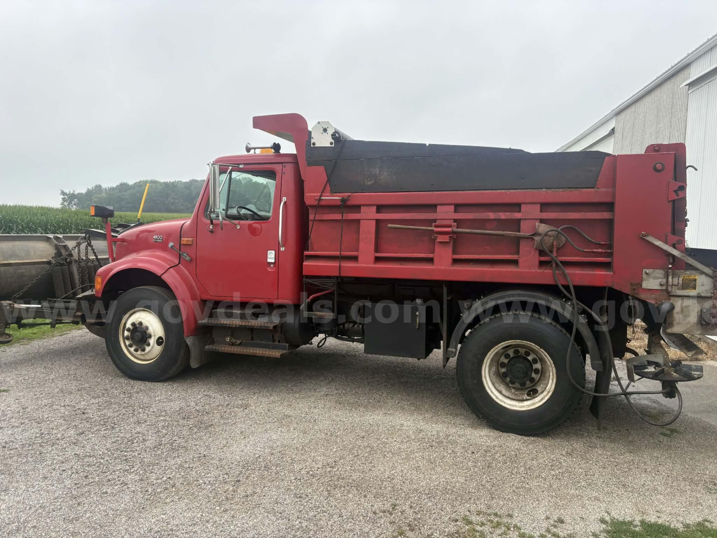 Dump truck with red cab and dump bed, side profile view showing large front wheel, black bed liner, and rear hydraulics. Visible text and stickers appear on body edges.