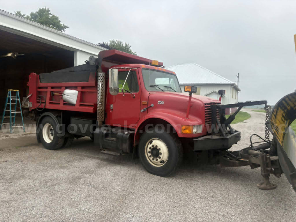 2000 International 4900 dump truck in red with front plow attachment, side dump bed, black grille, white wheels, parked near a building.