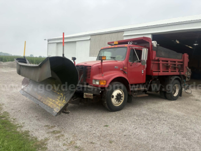 2000 International 4900 dump truck with snowplow or front plow attachment, red cab and dump bed, parked outside a large shed.