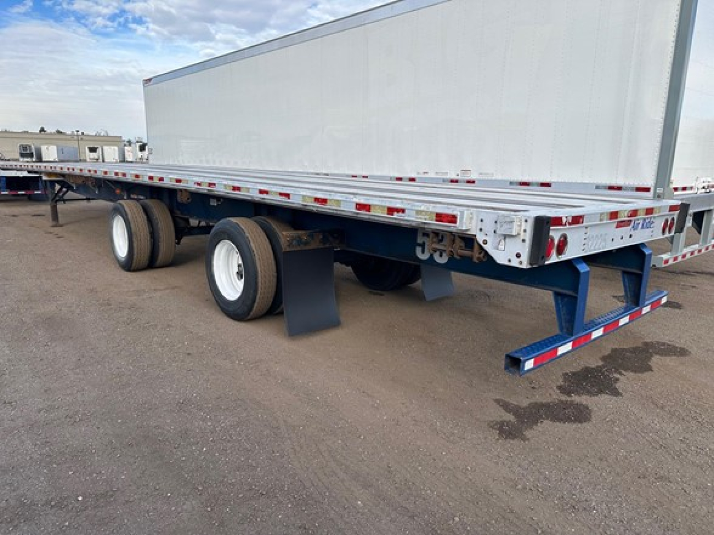 Flatbed trailer in side view showing dual rear axles, white wheels, and reflective safety tape along sides.